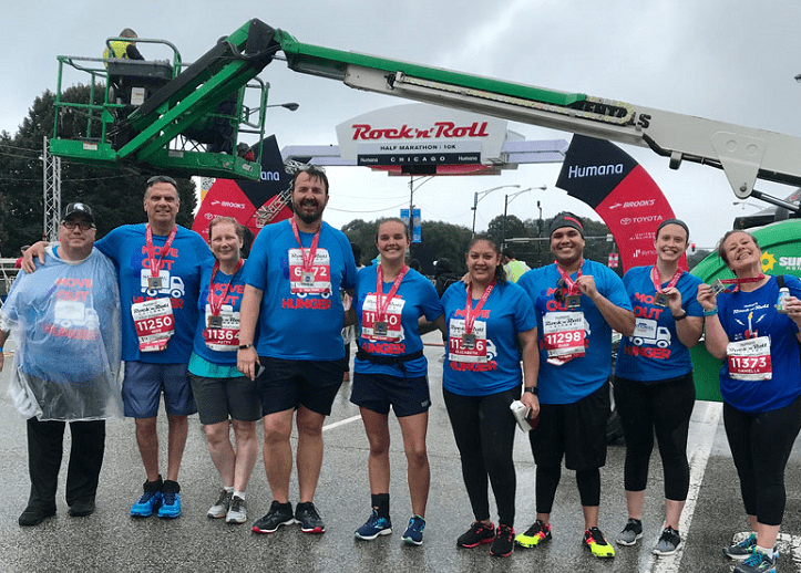 A group of nine people stands together, smiling and holding medals after completing a race. They are dressed in blue shirts and athletic wear. Behind them, an arch with signage reading 