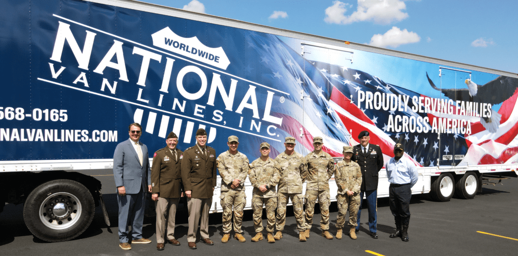 A group of eight people, including military personnel and civilians, stands in front of a large truck with a patriotic design featuring the American flag. The truck displays 